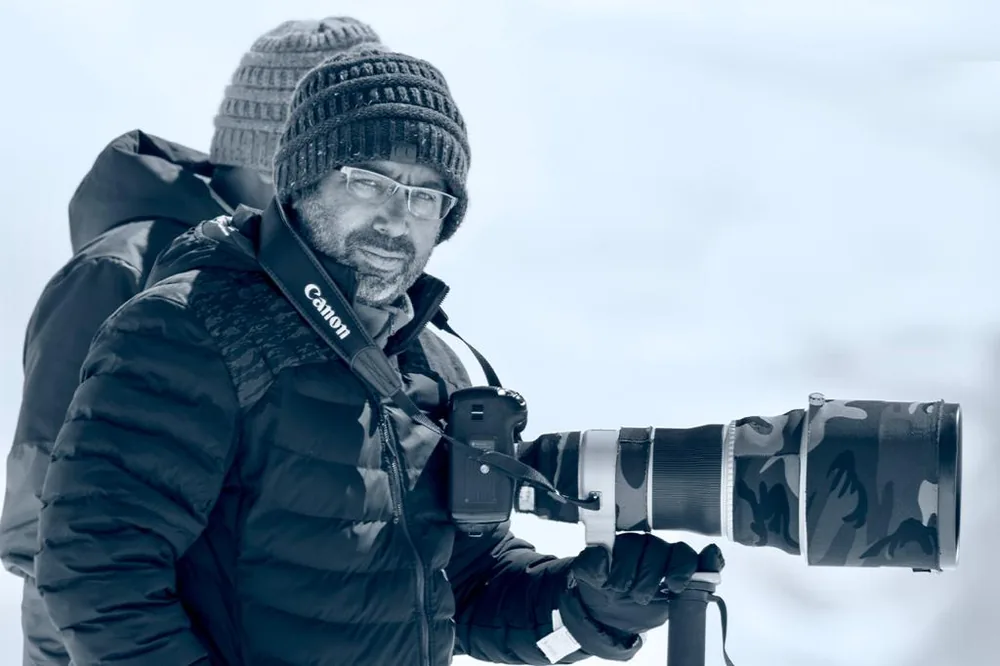 Vinit Arora in a woolen beanie and down jacket holding a camouflage-wrapped telephoto lens in a snowy winter landscape.